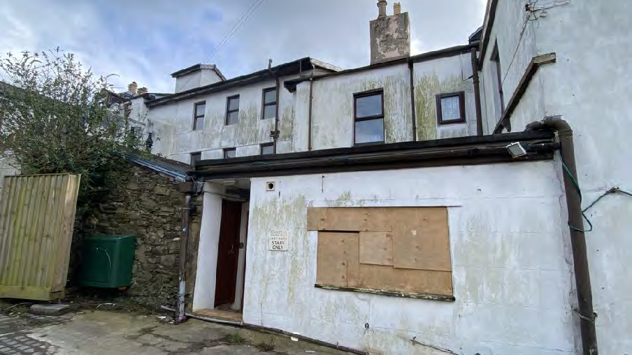A photograph showing the exterior of a weathered white building with a boarded-up window and a stone boundary wall.