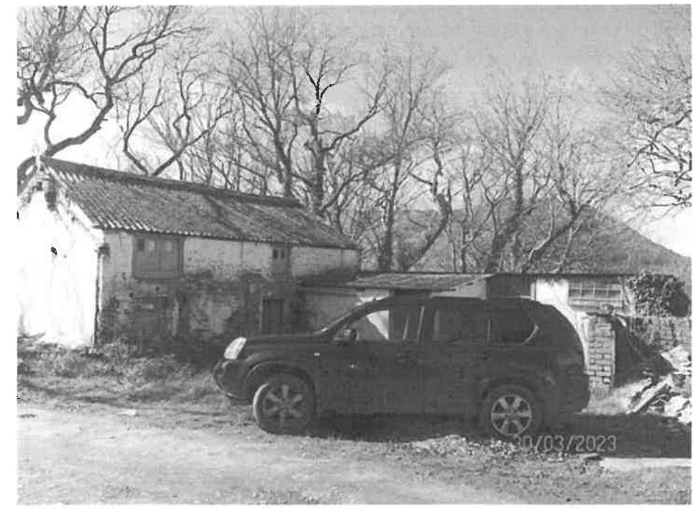 A black and white photograph showing an existing stone outbuilding or barn with a pitched roof, surrounded by trees and a parked SUV in the foreground.