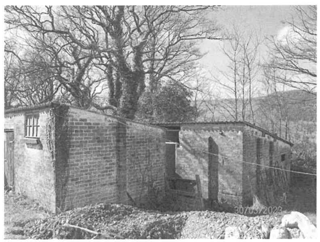 A black and white photograph showing a brick outbuilding or barn structure in a rural setting with trees in the background.