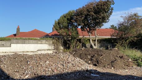 A photograph showing a construction site with large piles of rubble and earth in the foreground. An existing building with a red tiled roof is visible in the background behind trees and a concrete wall.