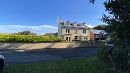 A photograph showing a large detached house with a pitched roof and dormer windows, set back from the road behind a hedge and driveway.