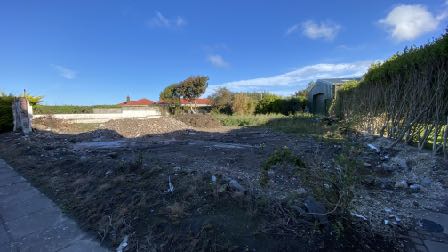 A photograph showing a cleared plot of land with piles of earth and rubble in the foreground, likely the site of the former dwelling. Existing houses and a tall hedge are visible in the background under a blue sky.
