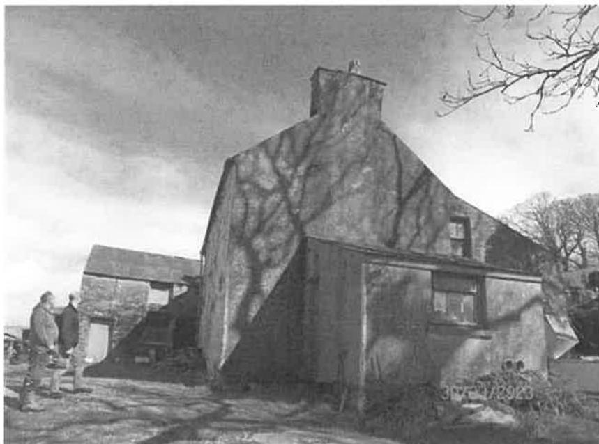A black and white photograph showing a large, old stone building, likely a barn, with two people standing nearby inspecting the exterior.