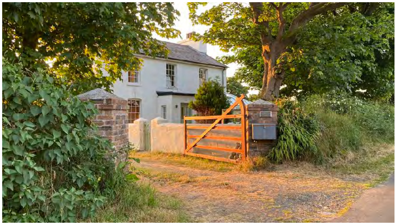 A photograph showing a white two-story detached house set back from the road behind a gravel driveway and wooden gate. The property is surrounded by mature trees and greenery, indicating a rural setting.