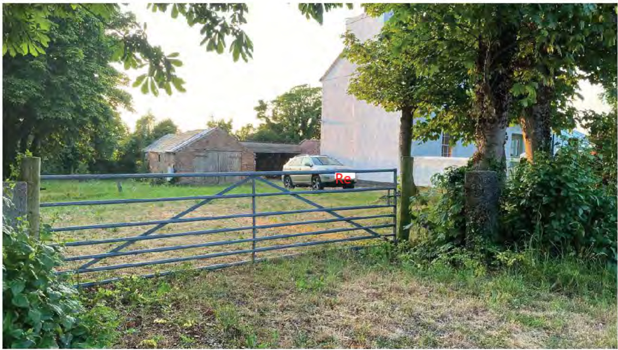 A photograph showing a rural property with a metal farm gate in the foreground. In the background, a brick outbuilding and a larger white building are visible behind the gate.