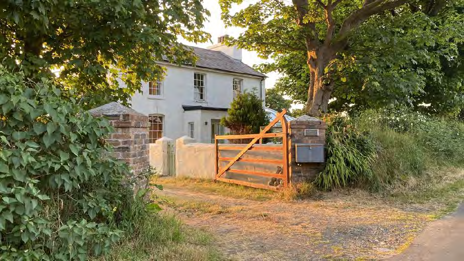 A photograph of a white two-story house with a slate roof, viewed through a wooden gate and stone pillars in a rural setting.