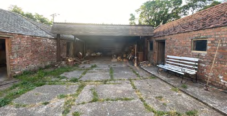 A photograph showing a rural courtyard surrounded by brick farm buildings, including a covered lean-to structure and a white bench.