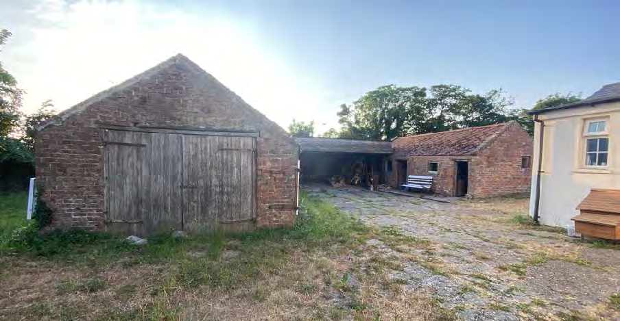 A photograph showing a large brick outbuilding with wooden doors, likely the former dairy barn proposed for conversion. The scene includes a gravel yard and adjacent rural structures.