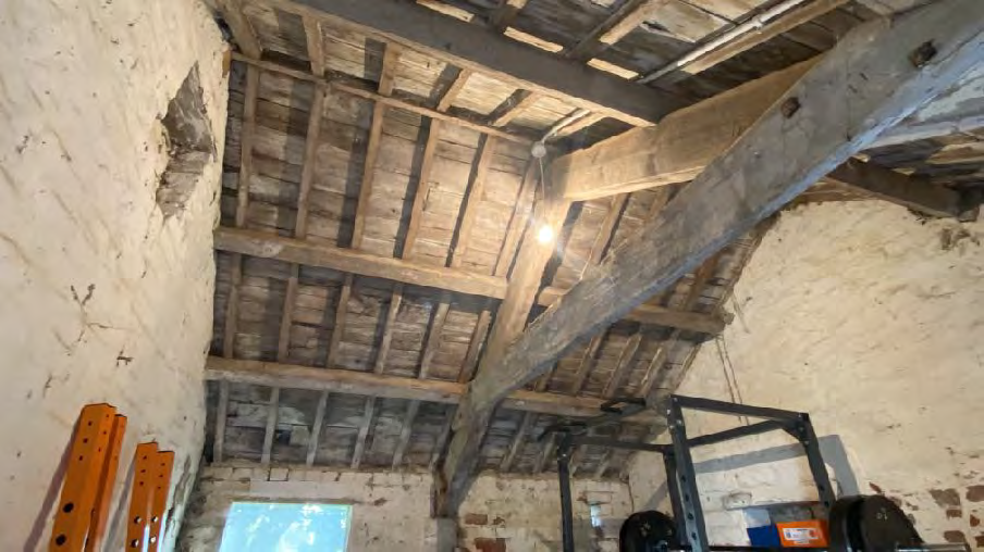 Interior photograph of a former dairy barn showing exposed wooden roof timbers, rafters, and plastered walls.