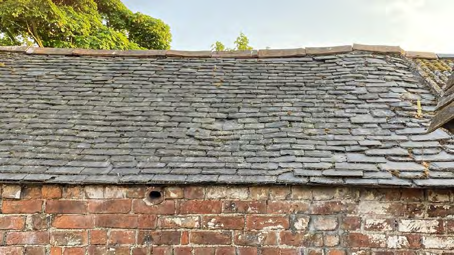 A close-up photograph showing the weathered grey slate roof and upper red brick wall of an existing building.