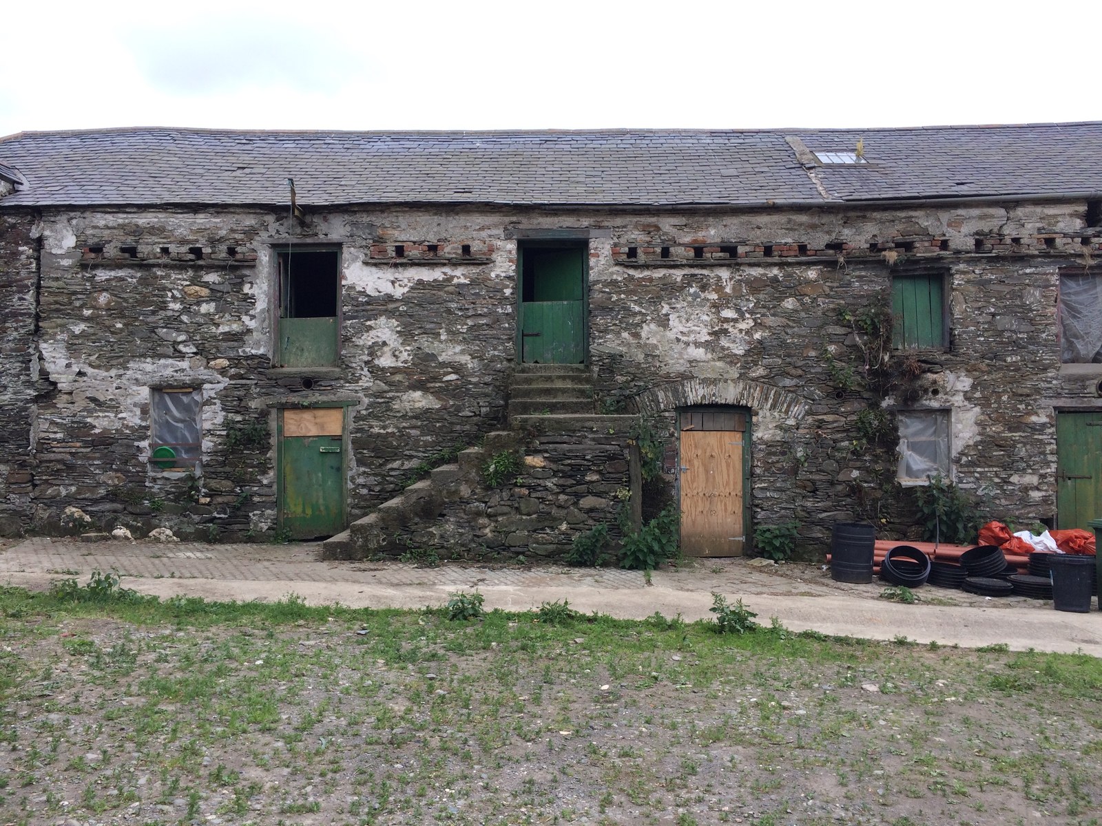 A photograph showing the existing facade of a long stone barn building with multiple door and window openings, some boarded up, and external stone steps leading to an upper-level entrance.