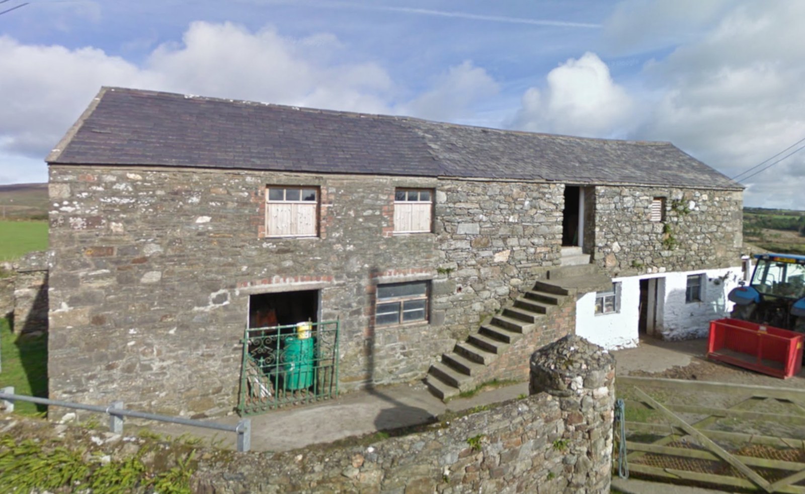 A photograph of a large, weathered stone barn with a slate roof, featuring stone steps leading to an upper entrance and a tractor parked nearby in a rural setting.