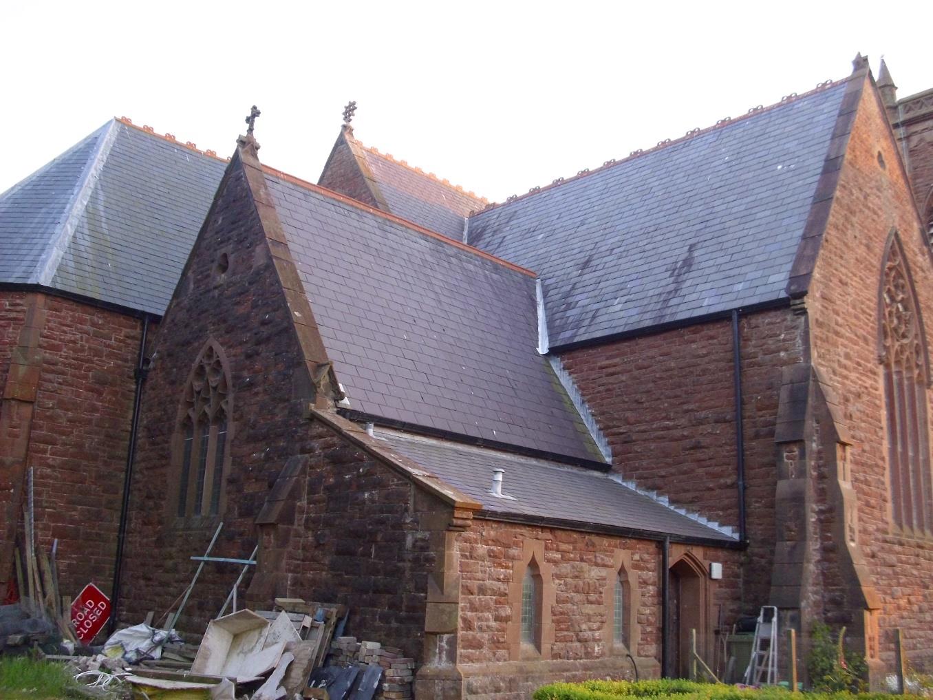 A photograph of a large red stone church or chapel with slate roofing, showing construction debris and a 'Road Closed' sign in the foreground.
