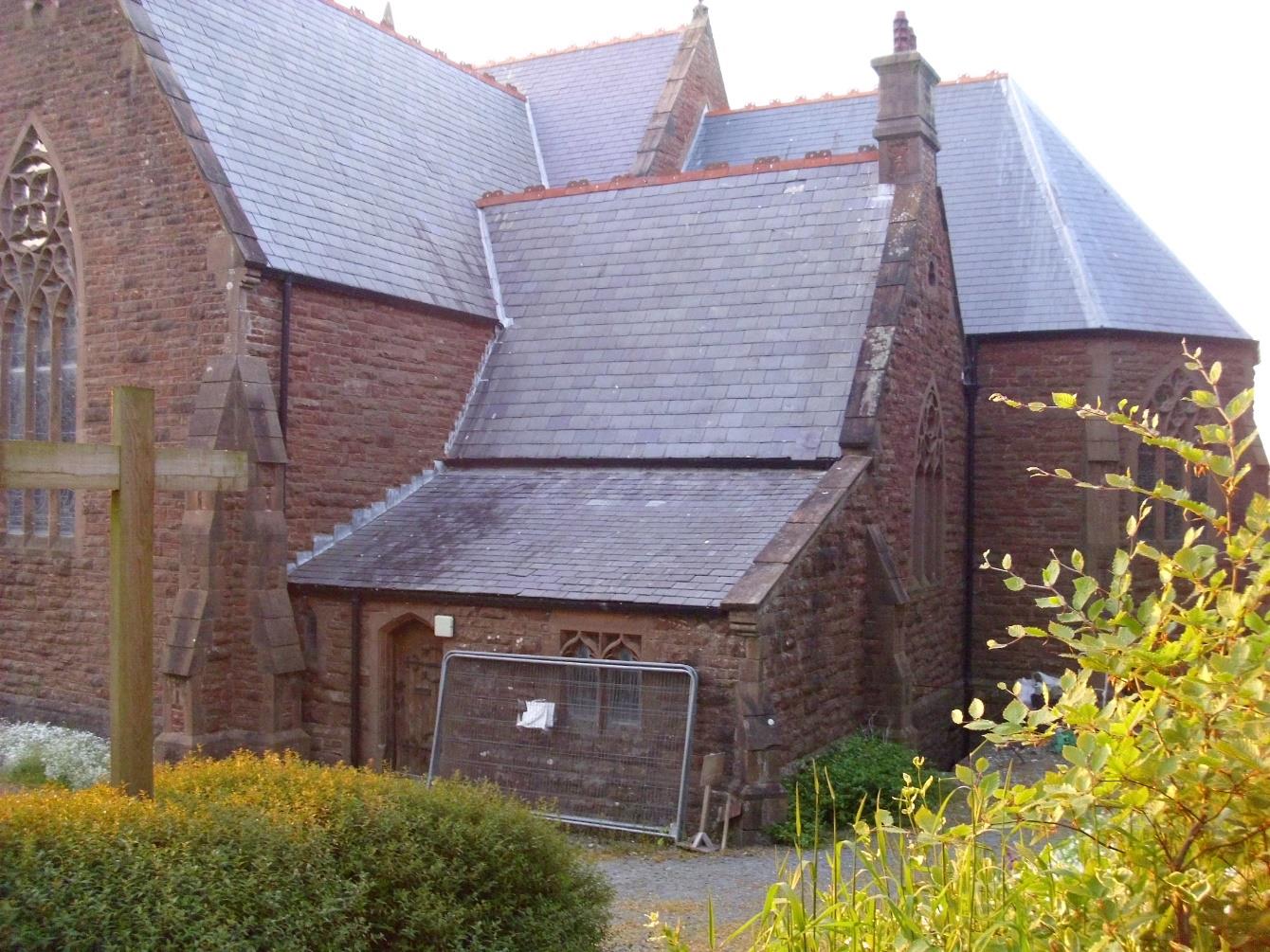 A photograph showing the exterior of a red brick church or chapel with Gothic-style windows and a slate roof, featuring a wooden cross in the foreground.