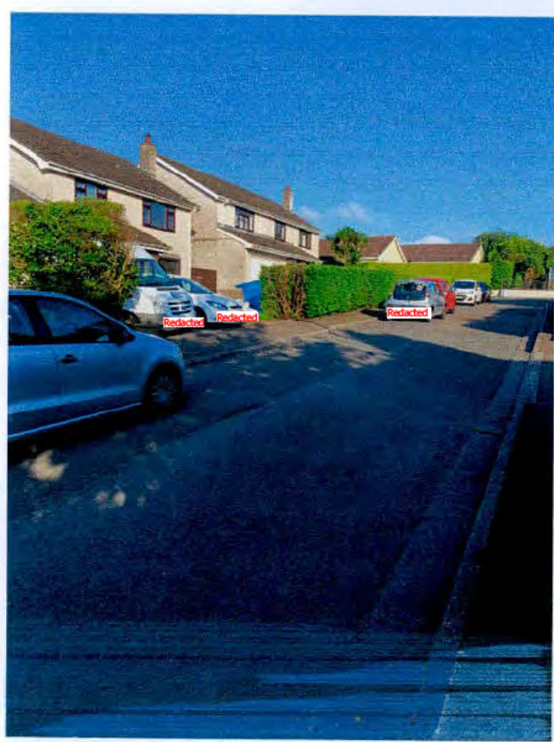 A street-level photograph showing a row of residential houses with cars parked along the curb and a green hedge.
