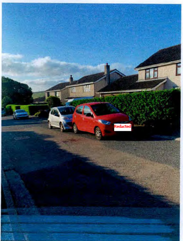 A photograph showing a residential street with parked cars and houses in the background.