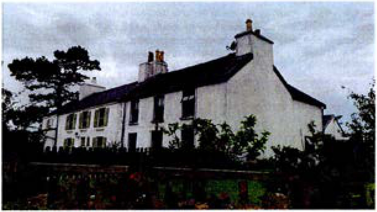 A low-resolution photograph showing a white, two-story residential building with a pitched roof and chimneys, surrounded by trees and vegetation.
