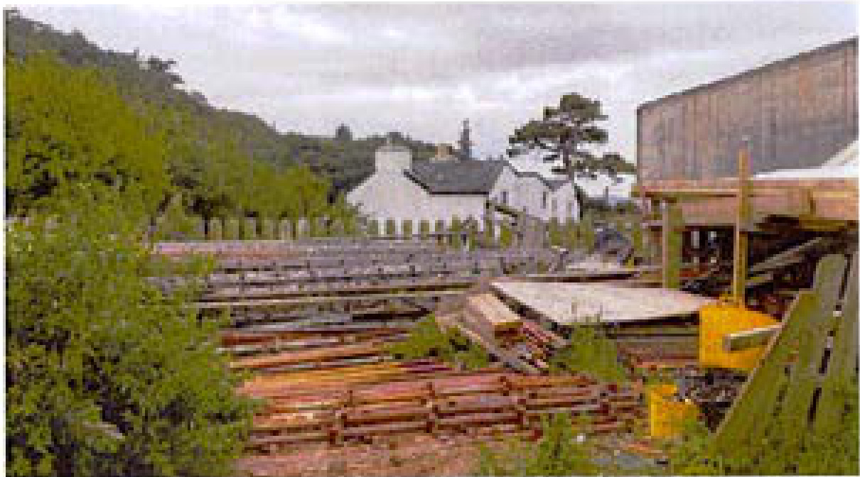 A photograph showing a yard stacked with timber and construction materials in the foreground, with white buildings and a hillside visible in the background.