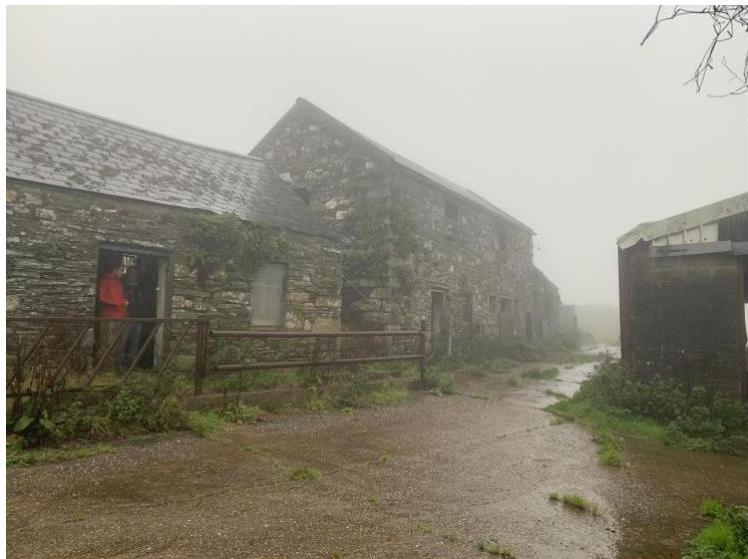A photograph showing a row of existing stone agricultural buildings or barns in a foggy rural setting, with a person visible in a doorway on the left.