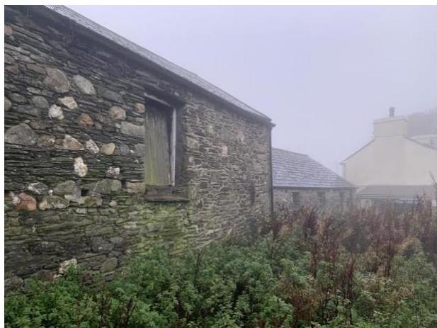 A photograph showing the exterior of an existing stone barn or outbuilding with a slate roof, surrounded by overgrown vegetation in a rural setting.