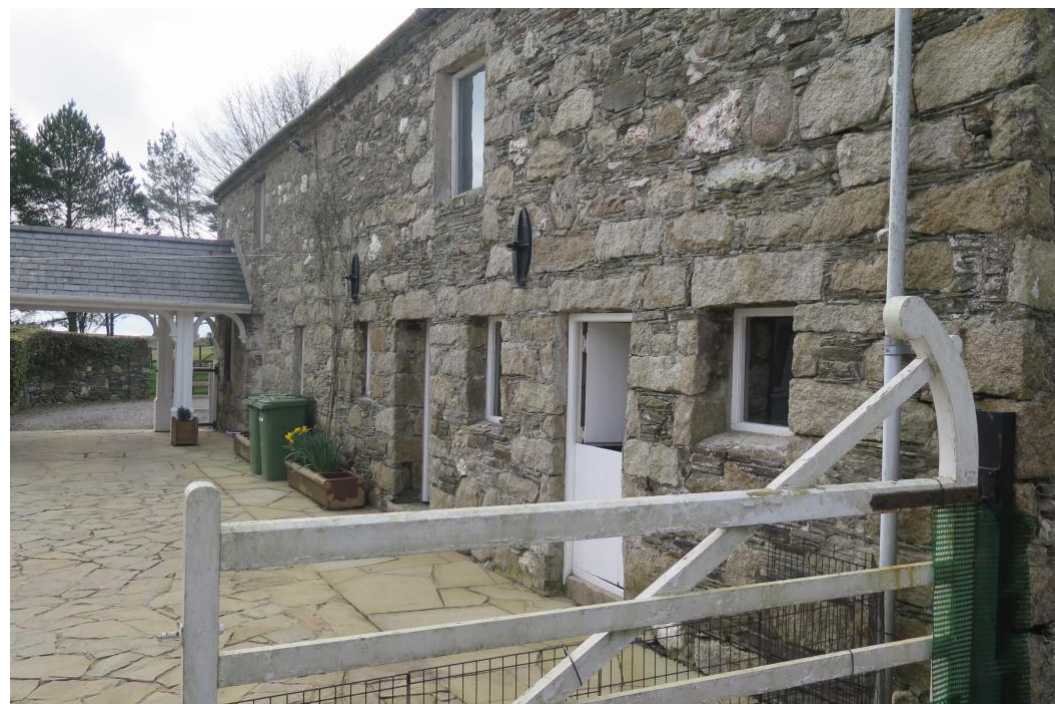 A photograph showing the exterior of a stone barn conversion with new windows and doors, featuring a covered link structure on the left and a white gate in the foreground.