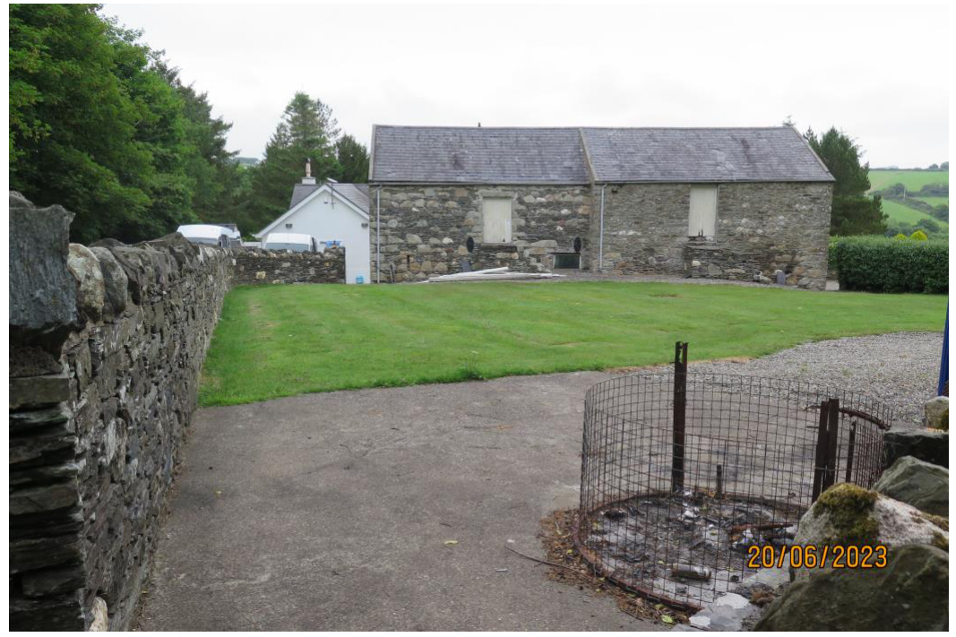 A photograph showing the rear garden, driveway, and existing stone barns of a rural property.