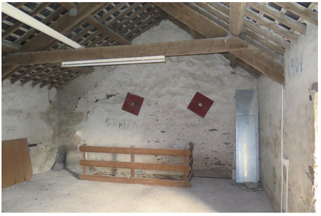 Interior photograph of a barn space showing exposed timber roof structure, roughcast walls, and a wooden fence-like structure.