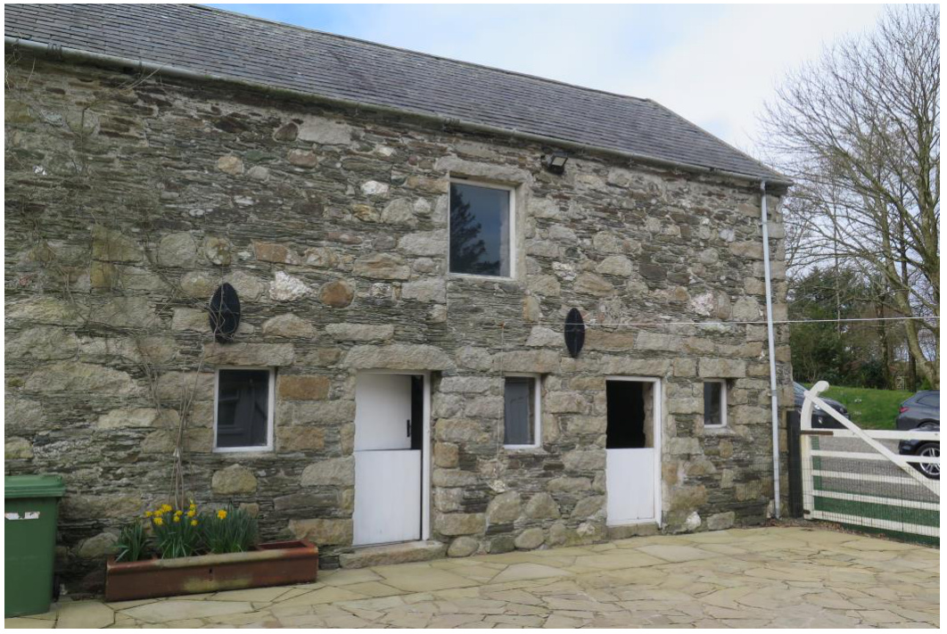 A photograph showing the exterior elevation of a stone barn or outbuilding with slate roofing, white doors, and a paved foreground.
