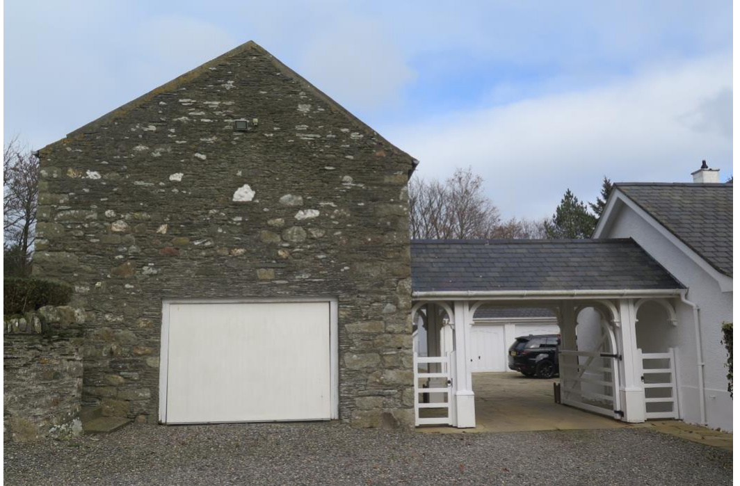 A photograph showing the existing site featuring a stone barn-like structure with a large white garage door adjacent to a white wooden carport and driveway.