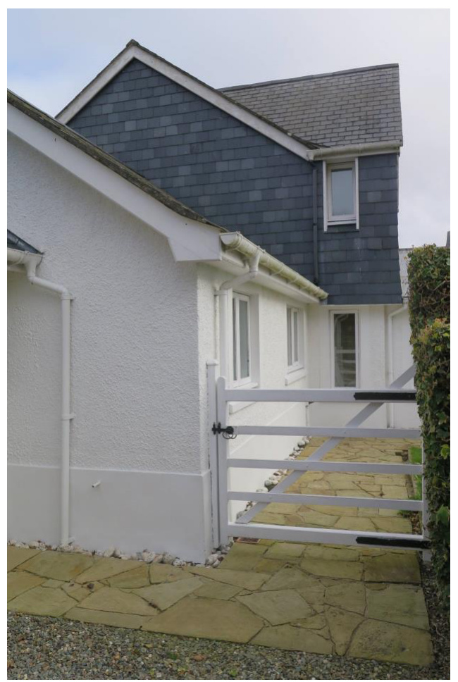 A photograph showing the side elevation of a white rendered building with a slate roof and a white gate leading to a paved path.