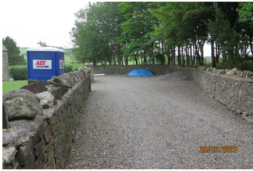 A photograph showing a gravel driveway area bordered by traditional stone walls, with a portable toilet visible on the left and trees in the background.