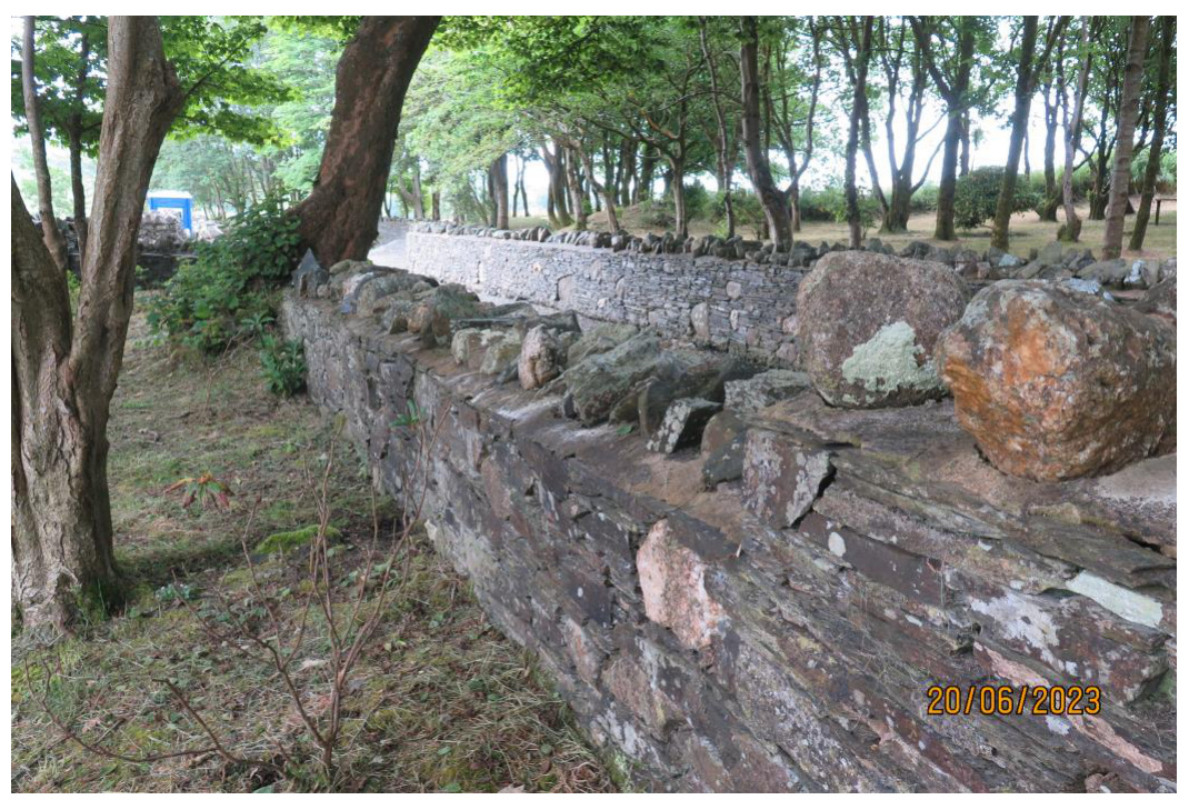 A photograph showing a long, dry-stone wall running through a wooded area with large boulders placed on top.