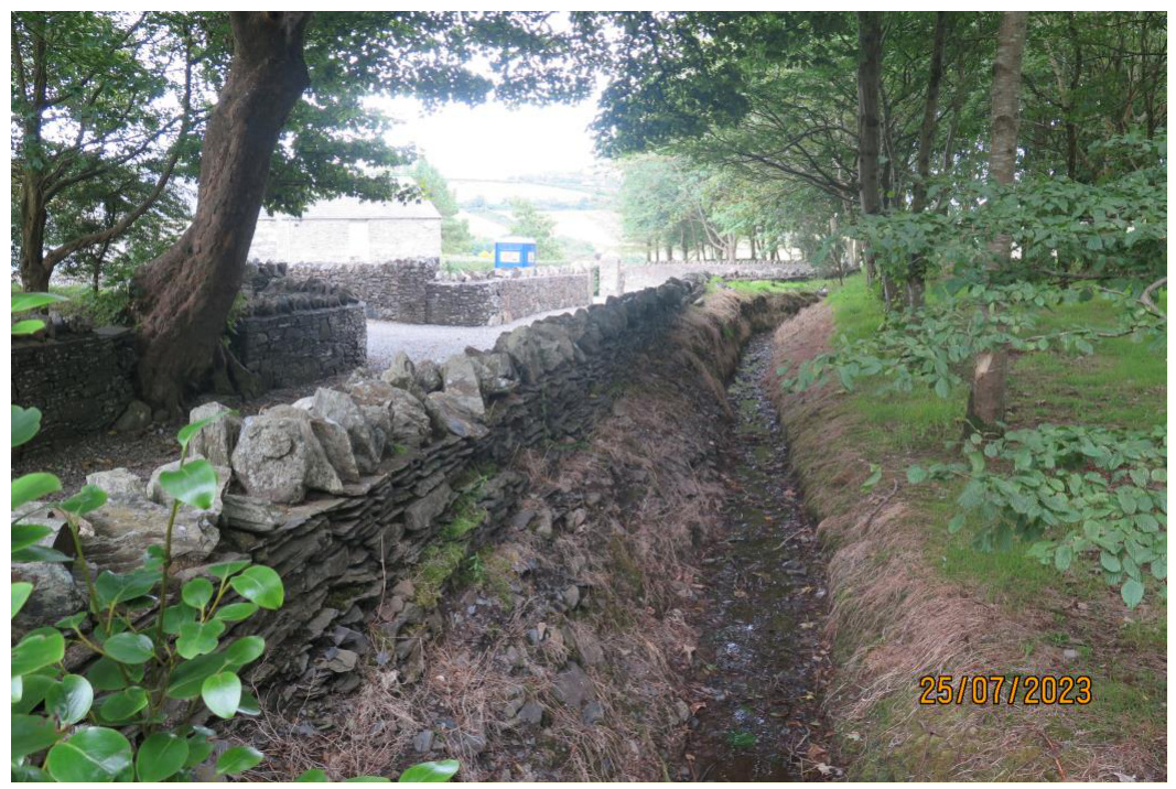 A photograph showing a rural site featuring a dry stone wall and a drainage ditch or trench running alongside it. A white building is visible in the background through the trees.