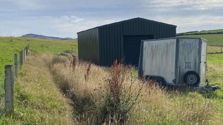 A photograph showing a large dark metal agricultural building, likely a stable block, situated in a grassy field with a horse trailer parked alongside it.