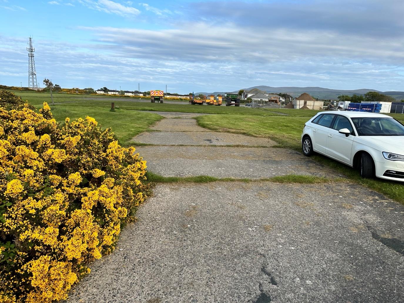 A site photograph showing a gravel access track leading into a grassy field with yellow gorse bushes in the foreground and construction vehicles in the distance.