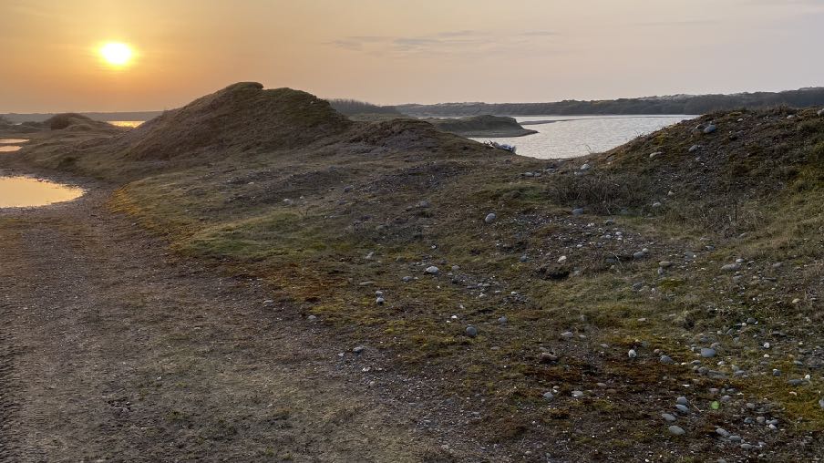 This photograph depicts a landscape view of a former quarry site featuring earth mounds, gravel paths, and small pools of water near a larger body of water. The scene captures the rugged terrain and natural surroundin...