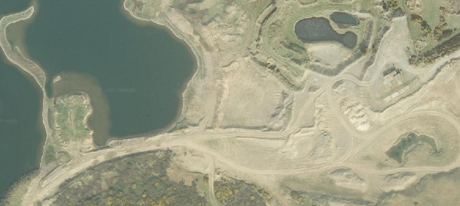 An aerial satellite view of a former quarry site adjacent to a large body of water, showing disturbed earth, access tracks, and small ponds.