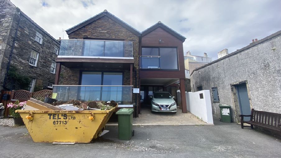 A photograph of a modern two-story detached house featuring stone and dark wood cladding, large glass windows, and a carport with a parked car. A large yellow skip sits in the foreground, indicating ongoing constructi...