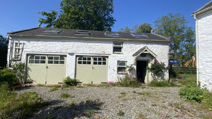 A photograph of a white stone building, likely a converted barn, featuring large garage doors, windows, and roof skylights in a rural setting.