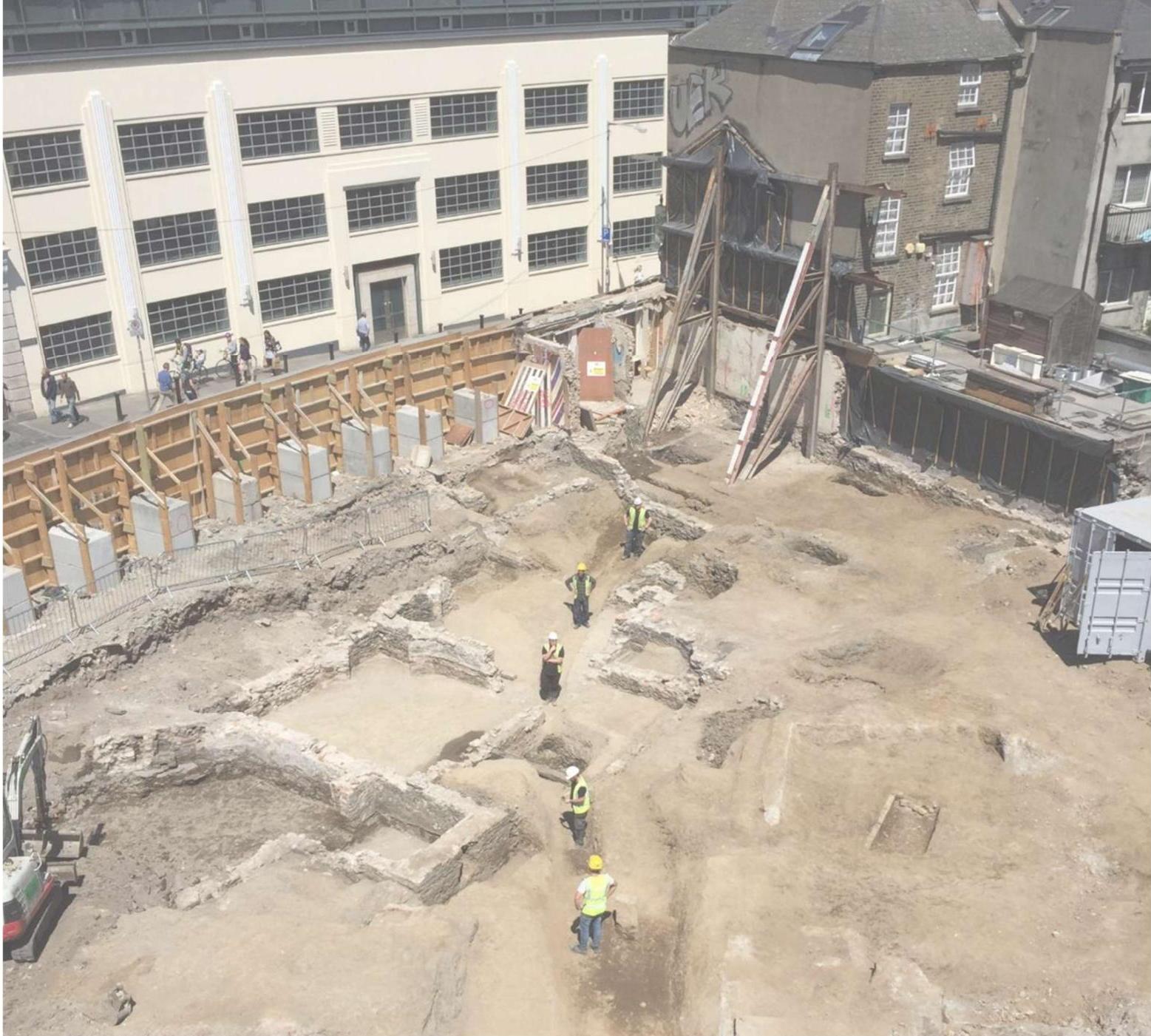 An aerial photograph showing an archaeological excavation site with exposed stone foundations and workers in high-visibility gear.