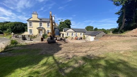 A photograph showing a rural property with a detached house and outbuildings, featuring an excavator on site indicating construction or reconstruction work.