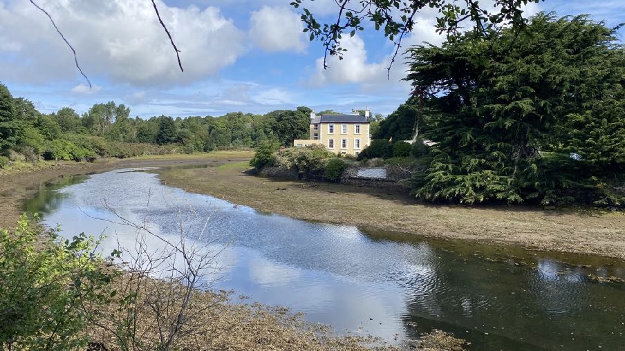 A photograph showing a large cream-colored detached house situated beside a river or estuary with muddy banks visible at low tide.