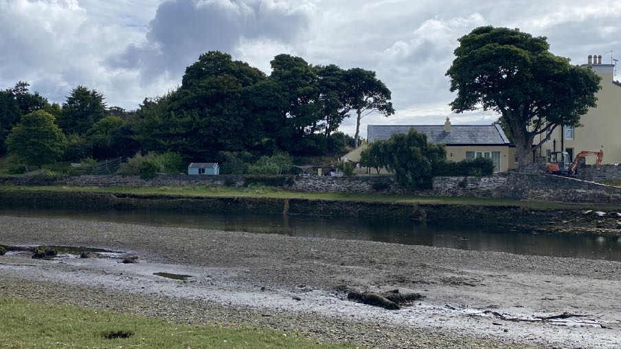 A photograph showing a tidal estuary in the foreground with a stone wall and residential buildings in the background.