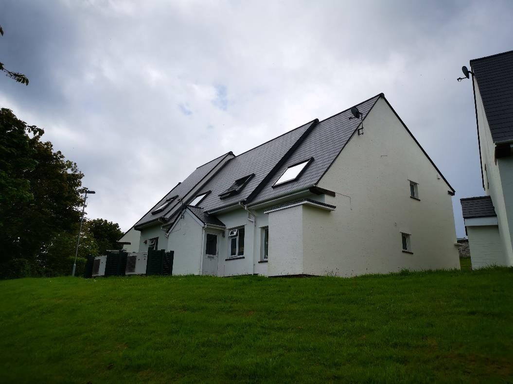 A low-angle photograph of a modern white detached house with a dark tiled roof and dormer windows, situated on a grassy slope.