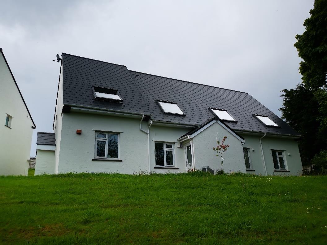A low-angle photograph of a single-story white detached house with a dark slate roof and multiple skylights, situated on a grassy slope.