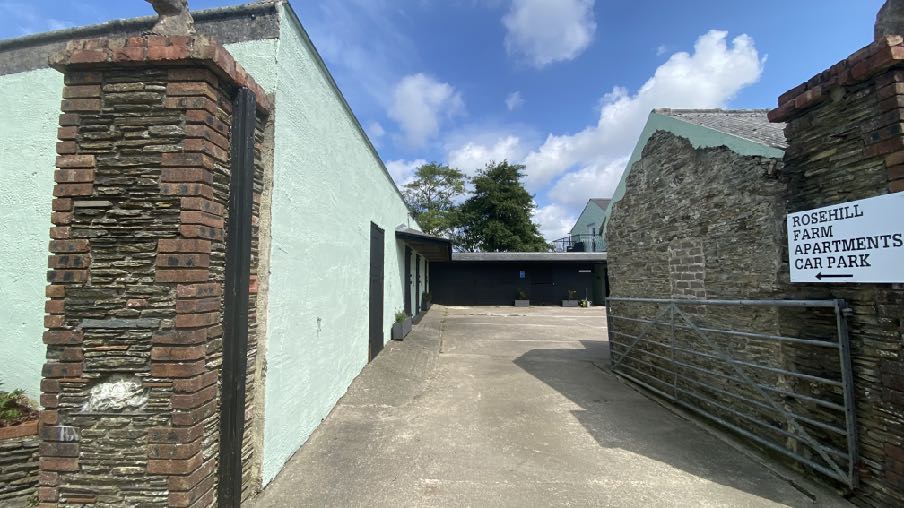 A photograph showing a paved access driveway and car park entrance between a rendered building and a stone wall, with a sign indicating 'Rosehill Farm Apartments Car Park'.