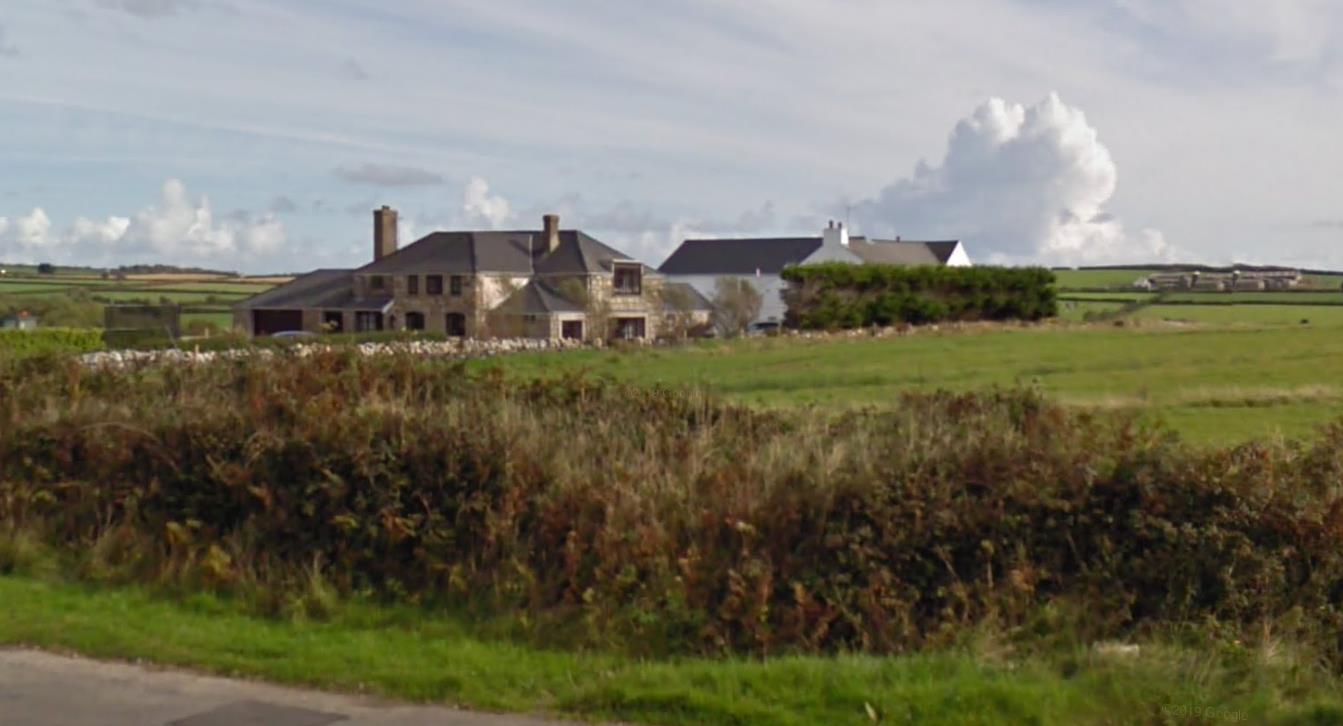 A photograph of a rural site featuring a stone house and a white building, likely farm structures, viewed from a roadside hedge.