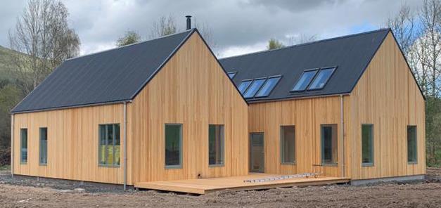 A photograph of a modern, timber-clad building with a dark pitched roof and skylights, situated on a construction site with bare earth and trees in the background.