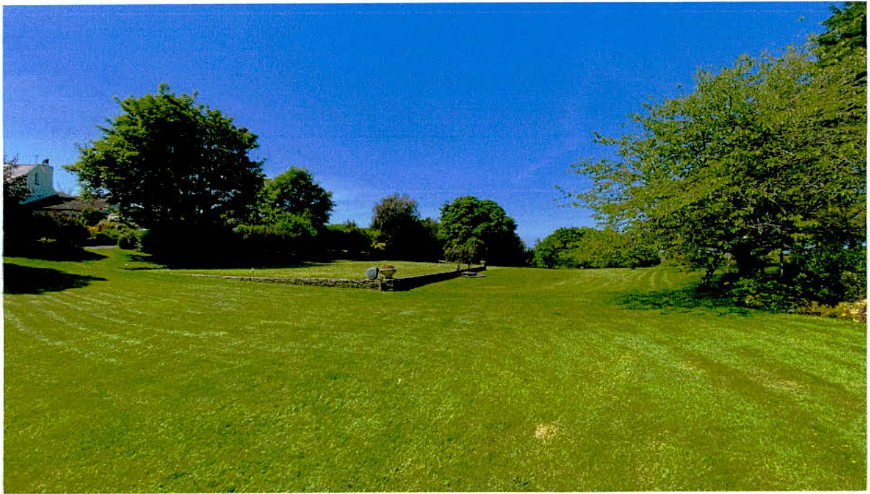 A wide-angle photograph showing a large, mown grassy lawn or field bordered by mature trees and a low stone wall. A portion of a white building is visible on the far left edge under a clear blue sky.