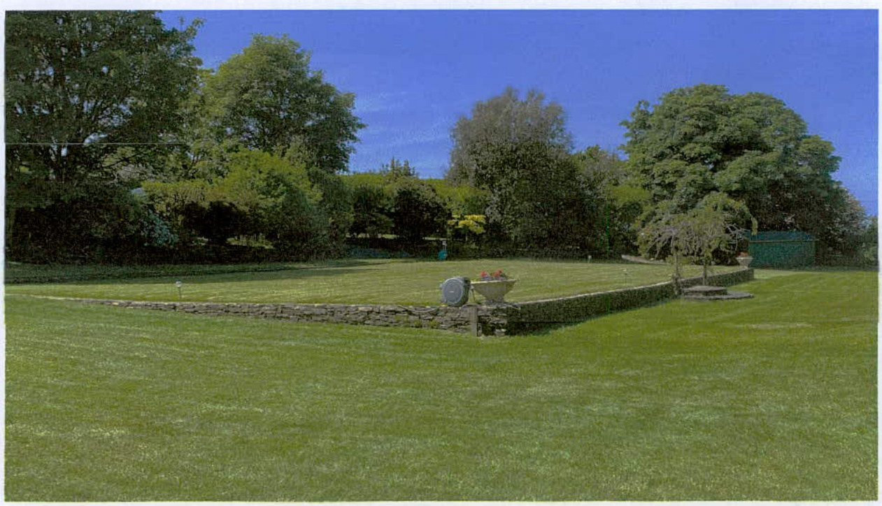 A photograph showing a grassy lawn area bordered by a low dry-stone wall with mature trees and a small green outbuilding in the background.